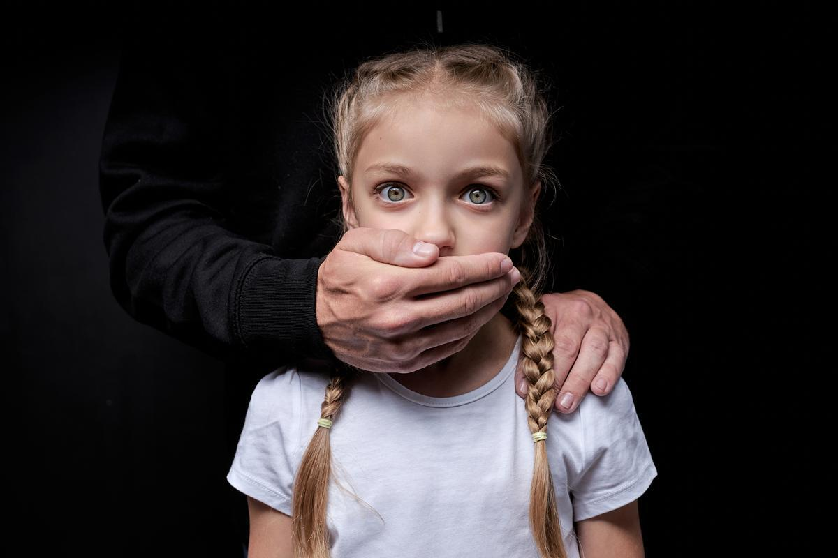 Child standing in front of an adult whose hand covers her mouth, illustrating coercion