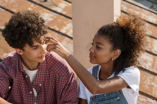 A teenage couple sitting close together outdoors, one gently touching the other's face