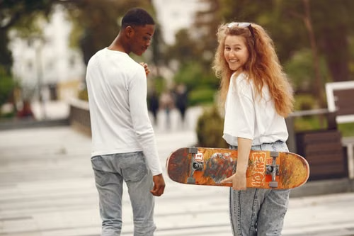 Young couple walking together in a city plaza with a skateboard, smiling at the camera